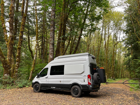 Light gray RV rental van with off-road tires, parked on a forest path amid moss-covered trees, ready for adventure.