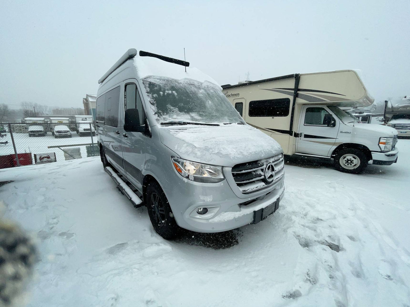  seen on a mountain road overlooking the sea and beach below.|Silver Mercedes Sprinter camper van & Class C RV covered in fresh snow at an RV rental lot. Perfect for winter adventures!|This silver camper van RV rental is perfectly positioned in a scenic mountain landscape with clear skies — photo 2