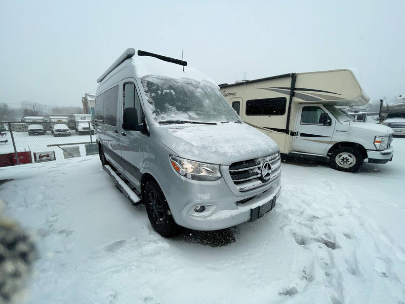  seen on a mountain road overlooking the sea and beach below.|Silver Mercedes Sprinter camper van & Class C RV covered in fresh snow at an RV rental lot. Perfect for winter adventures!|This silver camper van RV rental is perfectly positioned in a scenic mountain landscape with clear skies — photo 2