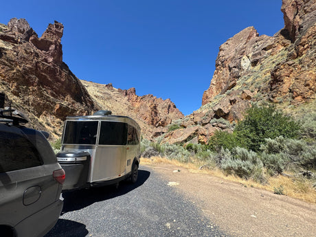 An Airstream rv rental and SUV parked in a dramatic canyon, surrounded by towering rock formations and clear blue sky.