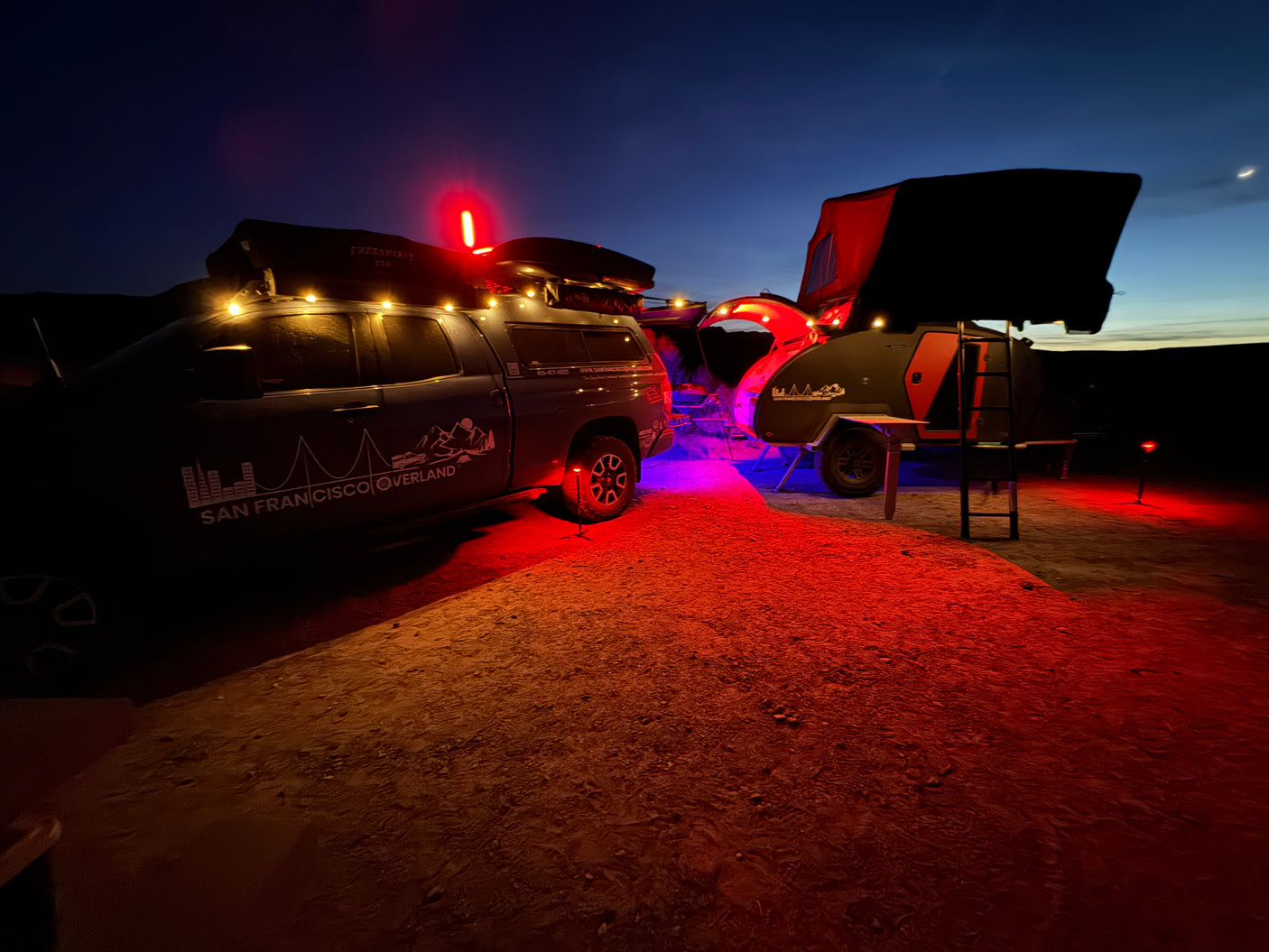 Illuminated night camp with "San Francisco Overland" truck & teardrop trailer under a crescent moon. RV rental ready!
