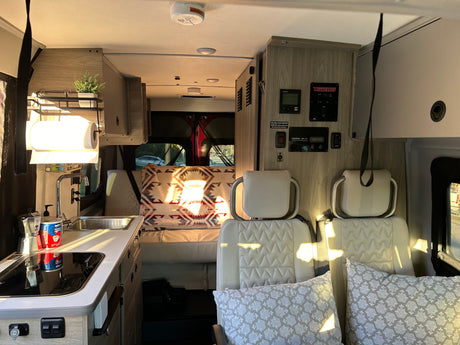 Inside a modern RV rental: a bright kitchenette with sink and stovetop, control panel, and comfortable seating leading to the living area.