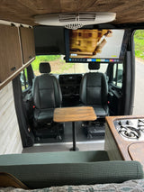 View inside an RV rental showing the front seats, a central table, an overhead TV, a kitchen counter with stove, and wooden cabinets.