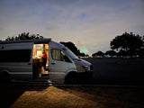 An RV rental provides a cozy space: A person cooks inside an illuminated camper van at night, with green fireworks in the distance.