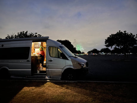An RV rental provides a cozy space: A person cooks inside an illuminated camper van at night, with green fireworks in the distance.