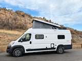 White Winnebago Solis camper van with pop-top extended, parked by rocky hills & dry grass under a cloudy sky. Perfect rv rental.
