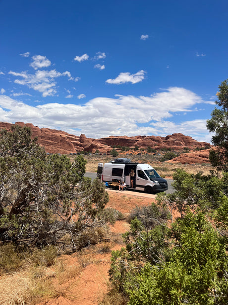 RV rental parked on desert road by red rock cliffs. Two people relax outside white camper van under blue, cloudy sky.