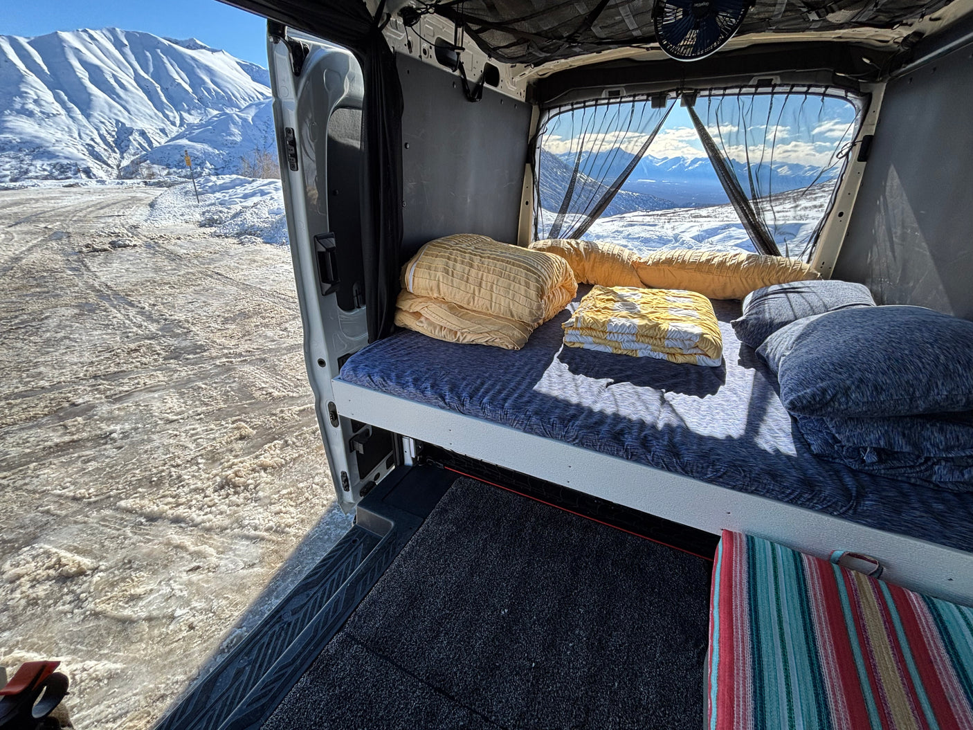 Step into this cozy RV rental bed, offering a panoramic view of snow-covered mountains and an icy winter road.