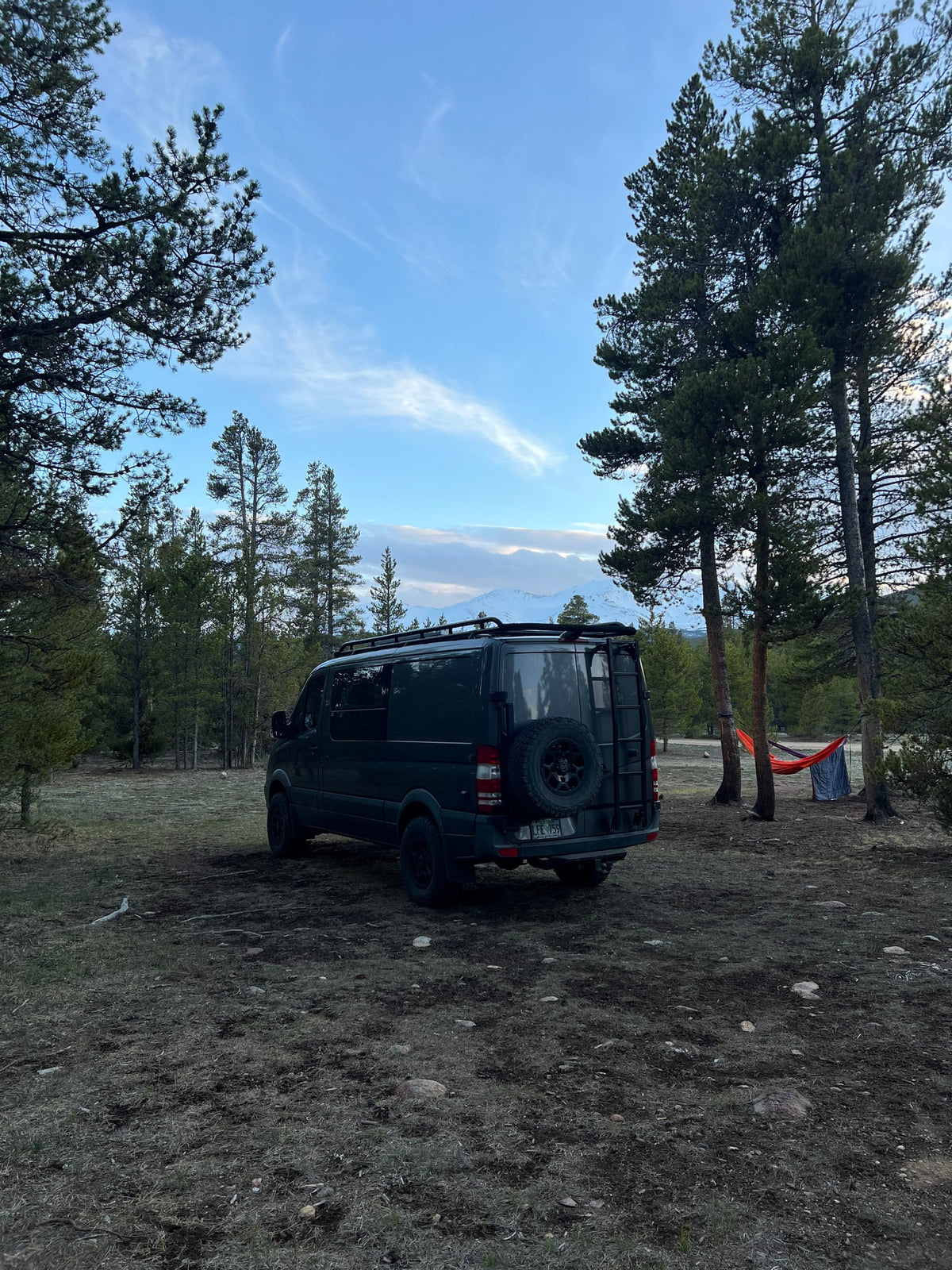 Dark RV rental van in a serene pine forest, with snowy mountains in the distance and a hammock for relaxation. — photo 6