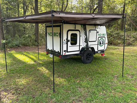 Explore nature! This small white camping trailer with an extended awning offers comfortable RV rental for outdoor travel in the woods.