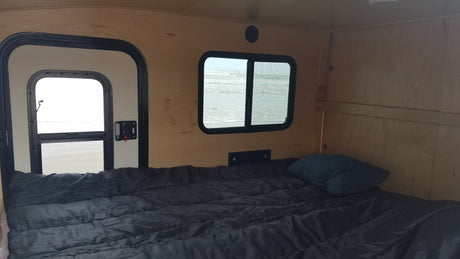 An RV rental's cozy bedroom features a dark comforter & pillow, with scenic views of ocean waves & beach through windows.