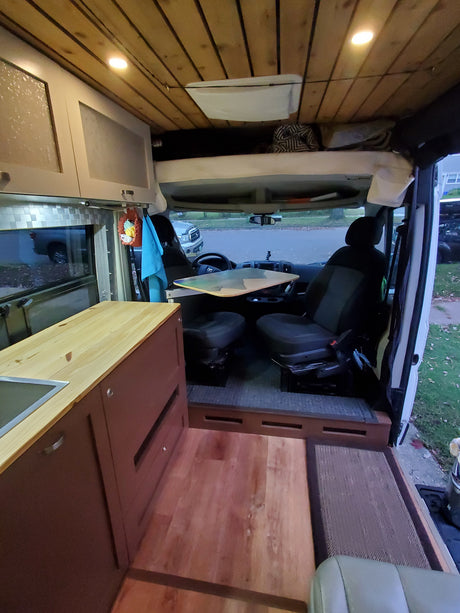Interior of a custom camper van conversion for RV rental. Features a kitchenette, front seats with a table, and elevated sleeping area.