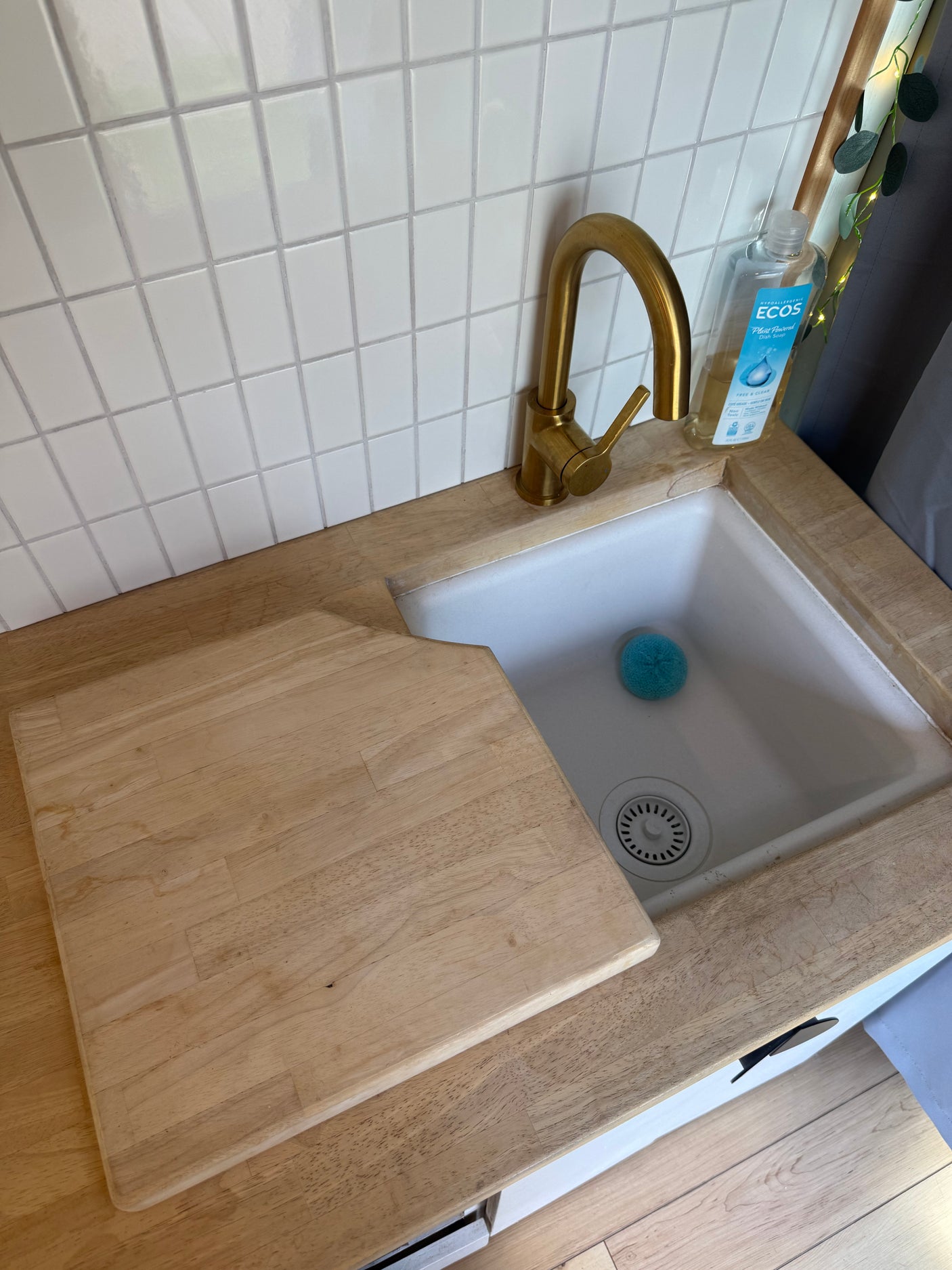 Compact rv rental kitchen featuring a white tiled backsplash, gold faucet, wooden countertop, and cutting board over the sink.
