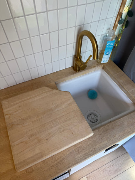 Compact rv rental kitchen featuring a white tiled backsplash, gold faucet, wooden countertop, and cutting board over the sink.