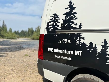White RV rental van with black tree graphics, parked by a river. WE adventure MT Van Rentals.