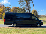 This black Mercedes-Benz Sprinter luxury van, an "Ultimate Coach," is ready for an rv rental, parked by grass and trees.