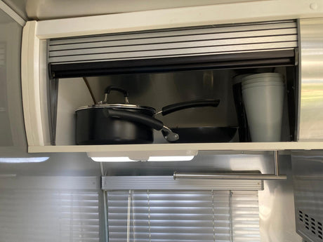 RV rental kitchen storage: An open overhead cabinet reveals black pots, a pan, and stacked gray cups. Under-cabinet light below.