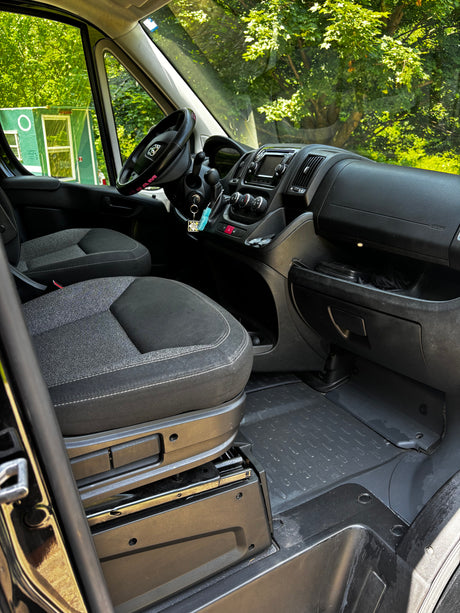 Interior of an RV rental van: clean, functional cabin with black and grey seats, dashboard, and a clear view of green trees.