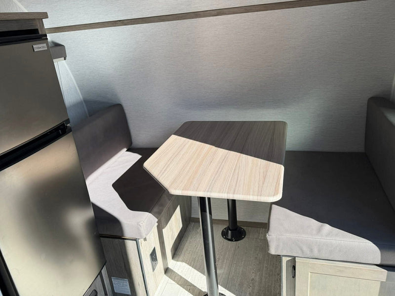 RV rental interior: dinette with light wood table, grey benches, and a refrigerator. Sunlight casts shadows. — photo 8