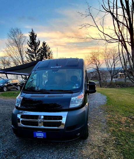 This black camper van RV rental sits on a gravel lot under a vibrant sunset sky, ready for your next trip.