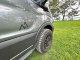 Adventure-ready gray RV rental van with an M1 Co. logo and rugged tires, parked on grass by a serene lake.