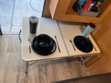 Compact dining in an RV rental: a folding table set with two black plates, silverware, and tumblers. View to outside.
