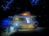 Night camping with an illuminated RV rental, pop-top open, string lights, and a portable washing machine at the campsite.