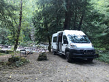 White camper van (rv rental) by a rocky river in a dense forest. Side door open, chair set up for riverside relaxation.