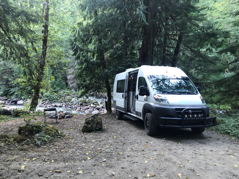 White camper van (rv rental) by a rocky river in a dense forest. Side door open, chair set up for riverside relaxation. — photo 13