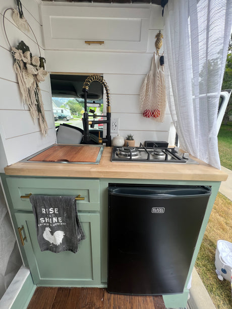 This RV rental offers a compact kitchen with sage green cabinets, a modern black and gold faucet, stovetop, and mini-fridge.
