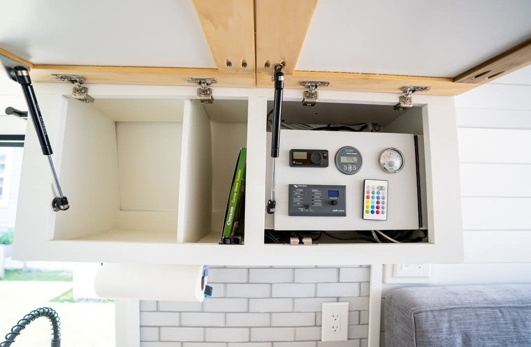 Open overhead cabinets in an RV rental reveal a power control panel, gauges, and a remote. Below are a paper towel holder & subway tile. — photo 20