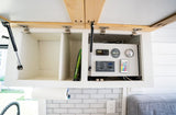 Open overhead cabinets in an RV rental reveal a power control panel, gauges, and a remote. Below are a paper towel holder & subway tile.