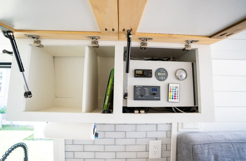 Open overhead cabinets in an RV rental reveal a power control panel, gauges, and a remote. Below are a paper towel holder & subway tile. — photo 20