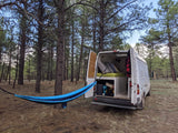 Relaxing campsite setup: a white camper van (RV rental) with open doors and a blue hammock in a peaceful pine forest.