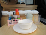 Compact RV rental kitchen with white dishes, mugs, and a pink crate of spices/cleaners, ready for adventure travel meals.