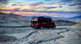 Adventure awaits! This maroon RV rental is parked on a vast, dry desert floor with eroding channels, mountains, and a vibrant sunset.