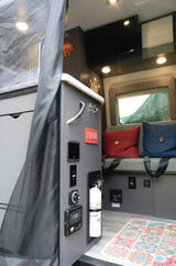Explore a comfortable RV rental interior with a control panel, fire extinguisher, cozy seating, and a scenic window view.