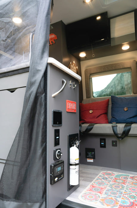 Explore a comfortable RV rental interior with a control panel, fire extinguisher, cozy seating, and a scenic window view.