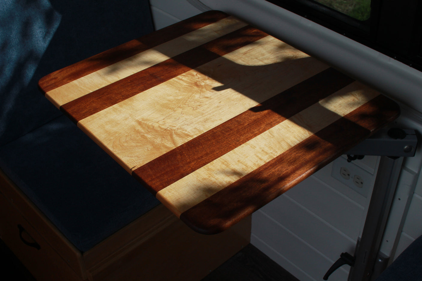 Striped wood table, illuminated by sun and shadows, in an RV rental. Note comfy seating & wall outlets for a versatile space.
