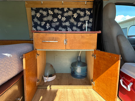 Inside this RV rental: functional kitchen with water pump, organized wooden storage, comfy bed, and a cooler for essentials.