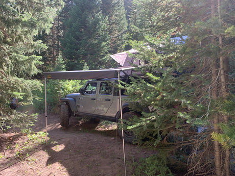 RV rental: Jeep Gladiator with roof tent & awning parked in a forest campsite, ready for adventure.