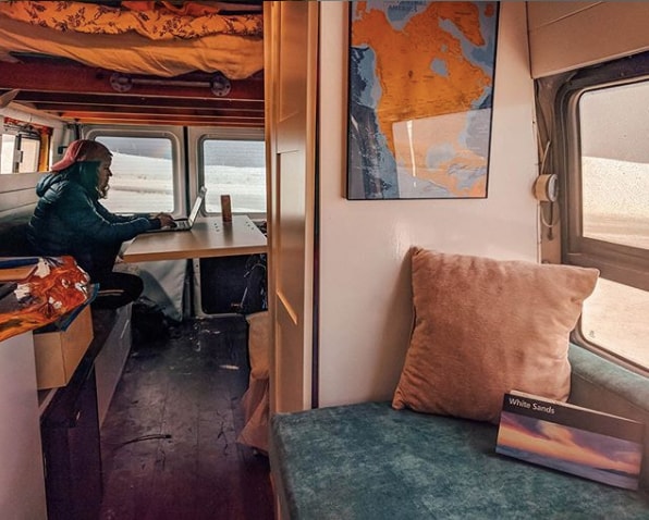 Experience remote work in a spacious RV rental. Person on laptop by window with White Sands view. Van interior with map, comfy seat.