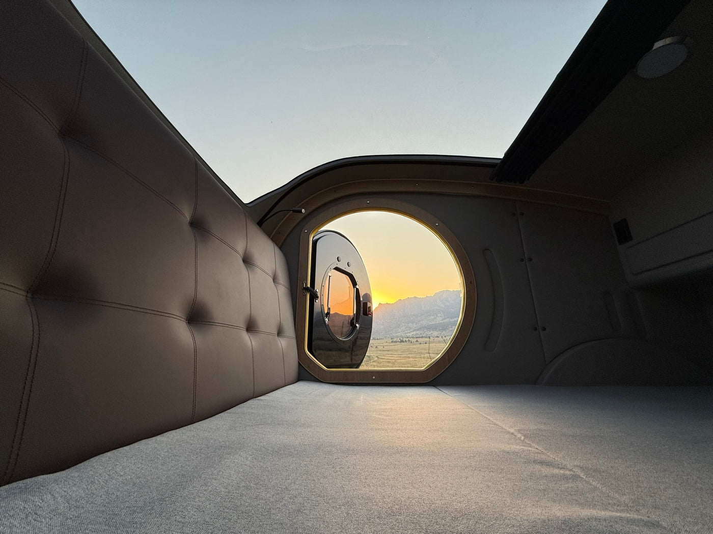 Comfortable rv rental interior: Tufted bed, unique round window open to a vibrant mountain sunset. Perfect for adventure. — photo 9