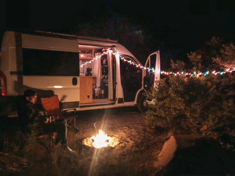 Person relaxing by a bright campfire next to a lit RV rental at night, adorned with colorful string lights.