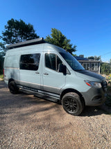 A modern gray Storyteller Overland adventure van with black wheels, parked on gravel. Consider this RV rental for your next trip.
