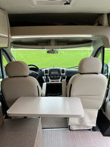 Interior of a modern **RV rental**, showing front seats, table, dashboard, and a green field through the windshield.