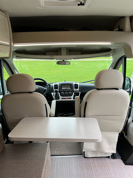 Interior of a modern **RV rental**, showing front seats, table, dashboard, and a green field through the windshield.