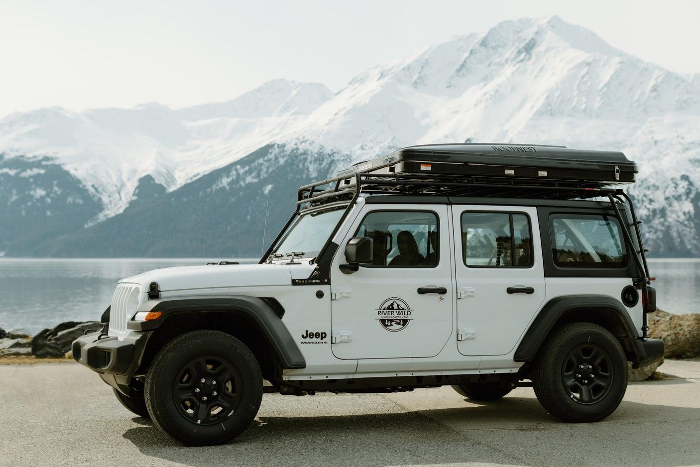 White Jeep Wrangler RV rental with rooftop tent by an Alaskan lake. Explore snowy mountains & wild landscapes.