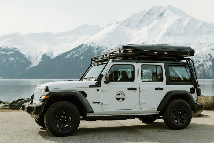 White Jeep Wrangler RV rental with rooftop tent by an Alaskan lake. Explore snowy mountains & wild landscapes.