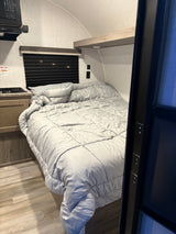 RV rental interior: A compact bedroom with a grey bedspread, small kitchen counter, stove, microwave, and wood flooring.